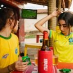 Mulher sorridente vestindo uma camisa de time tailandesa feminina da Seleção Brasileira, celebrando um gol em um estádio de futebol.