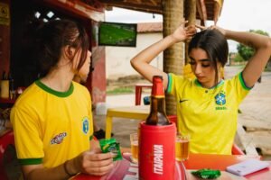 Mulher sorridente vestindo uma camisa de time tailandesa feminina da Seleção Brasileira, celebrando um gol em um estádio de futebol.