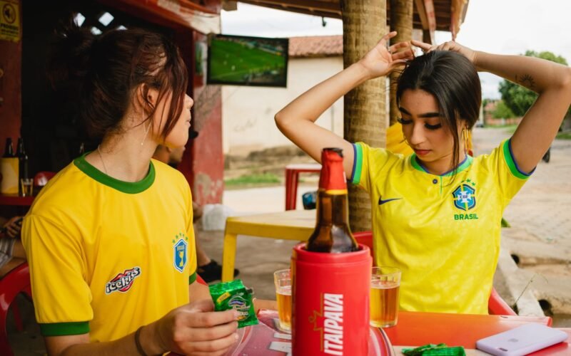 Mulher sorridente vestindo uma camisa de time tailandesa feminina da Seleção Brasileira, celebrando um gol em um estádio de futebol.