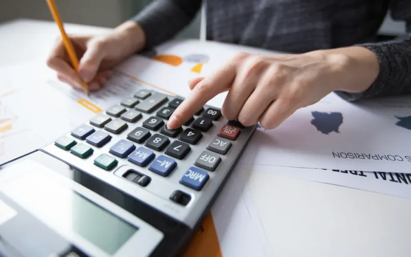 Pessoa revisando documentos financeiros e gráficos em um computador, com uma calculadora e caneta na mesa, simbolizando precisão e auditoria contábil.