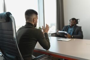 Pessoa sorrindo e gesticulando em uma entrevista de emprego, com um entrevistador atento e notas em espanhol.