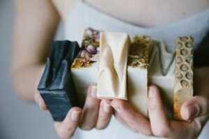 Mãos segurando um sabonete artesanal perfumado com flores secas, ao lado de um notebook com gráficos de vendas online.